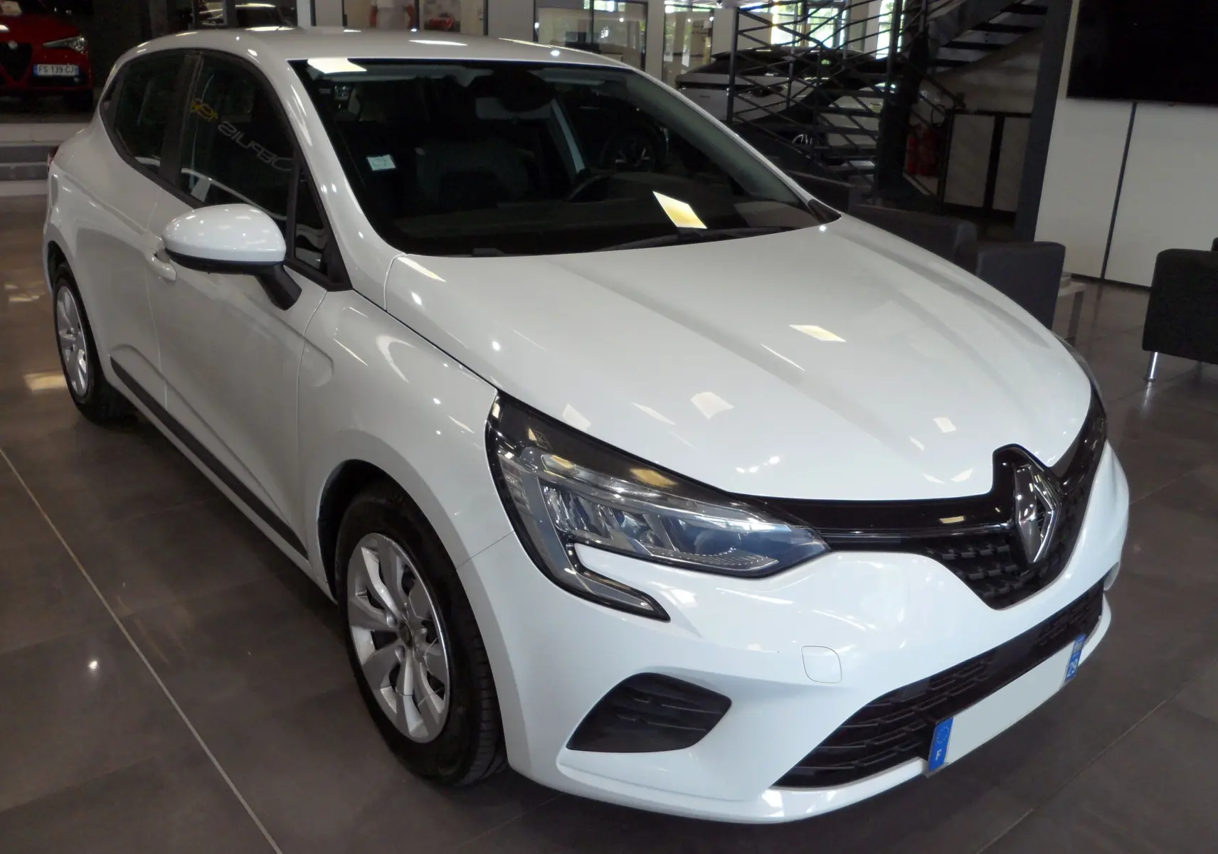 Renault Clio V blanche Glacier vue 3/4 avant droit en showroom, avec phares LED et calandre noire brillante.