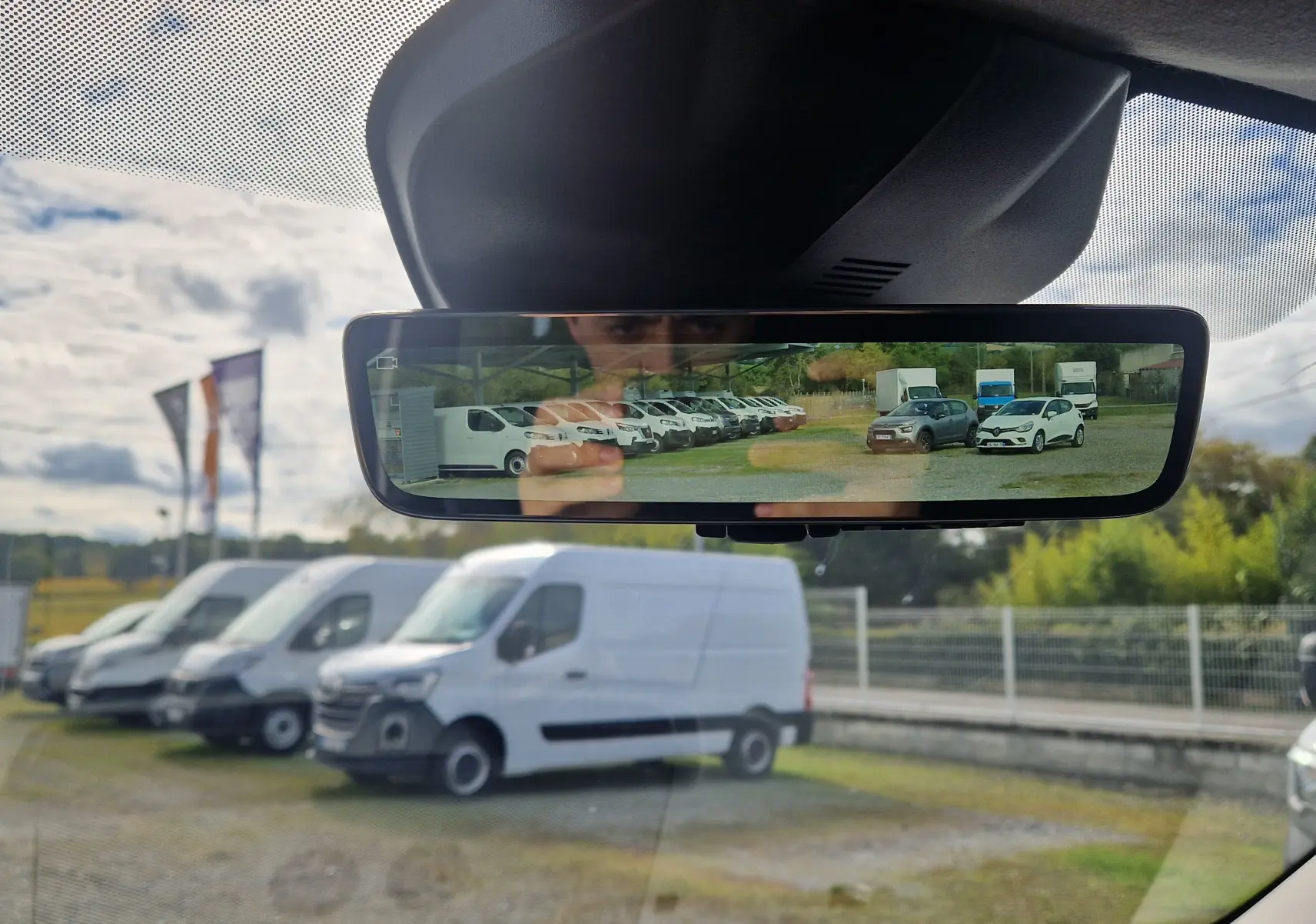 Rétroviseur intérieur montrant plusieurs véhicules utilitaires blancs en stationnement, avec reflet clair et ciel nuageux.