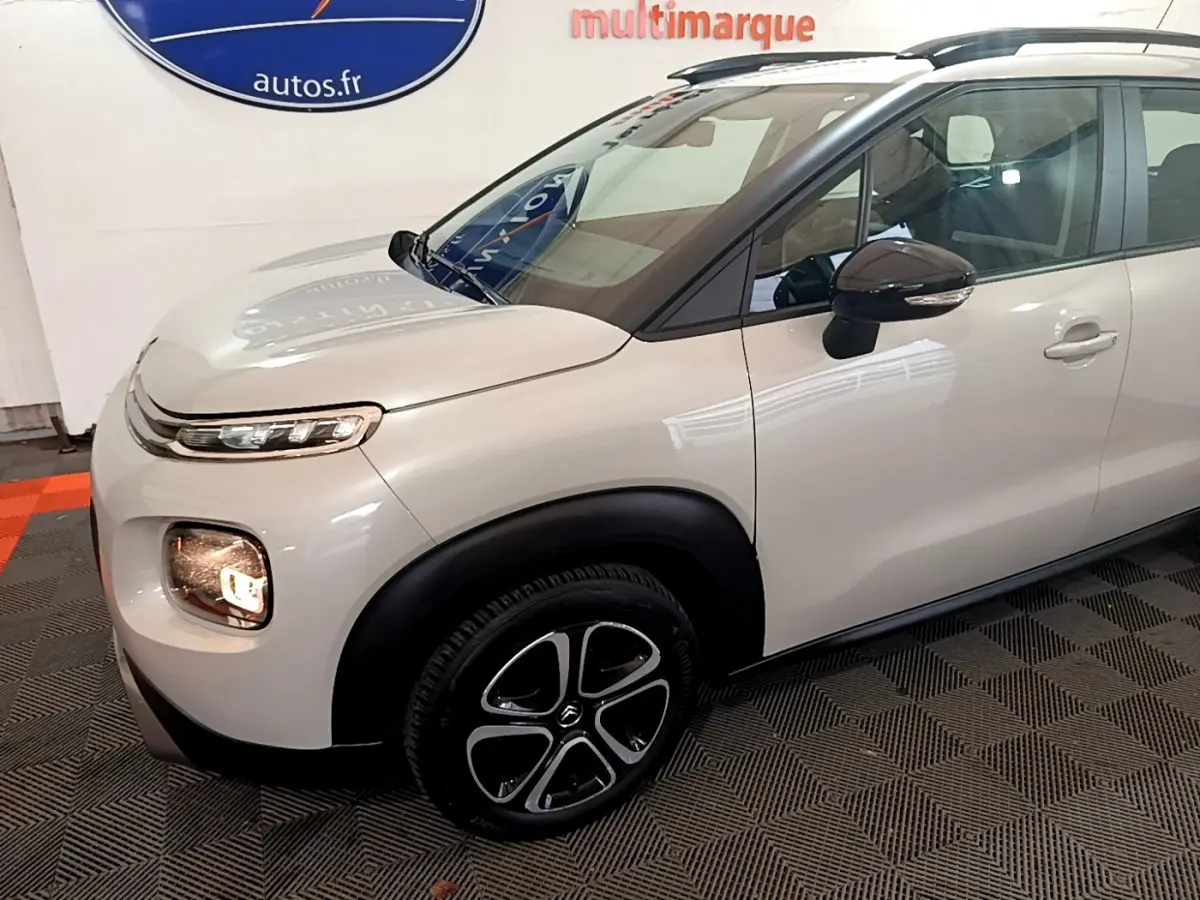 Vue 3/4 avant droit d'un Citroën C3 Aircross blanc avec jantes noires et barres de toit noires.