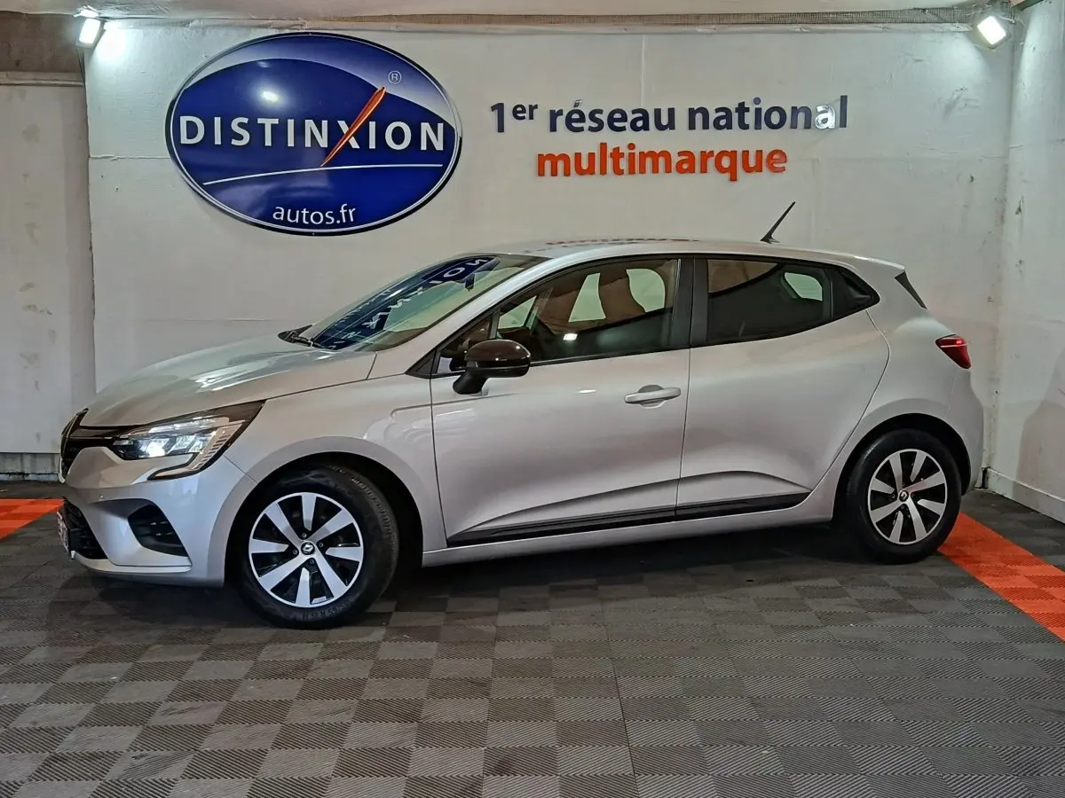 Profil droit d'une Renault Clio 2023 gris clair, avec jantes bicolores et rétroviseurs noirs, en intérieur showroom.