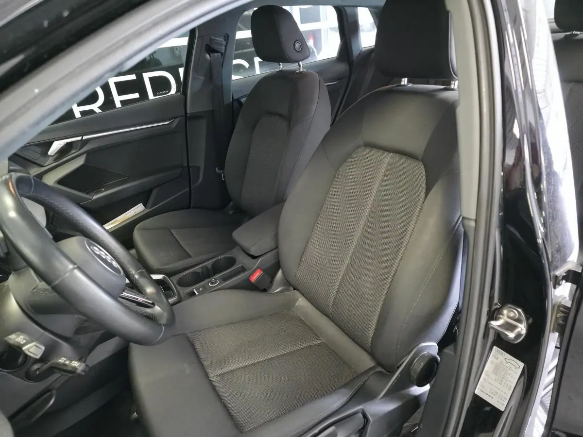Vue intérieure côté conducteur de l'Audi A3 Sportback 2023 noire, montrant les sièges tissu et le volant cuir multifonctions.