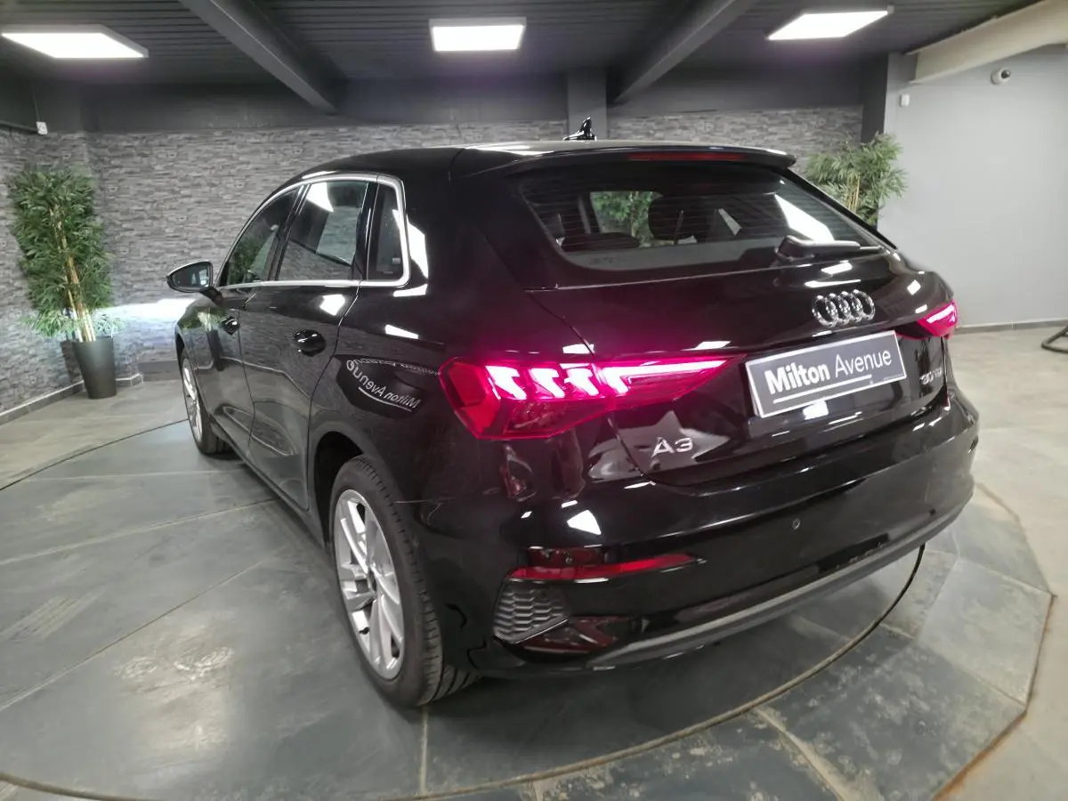 Audi A3 Sportback noir vue 3/4 arrière droit avec feux arrière LED allumés en intérieur showroom.