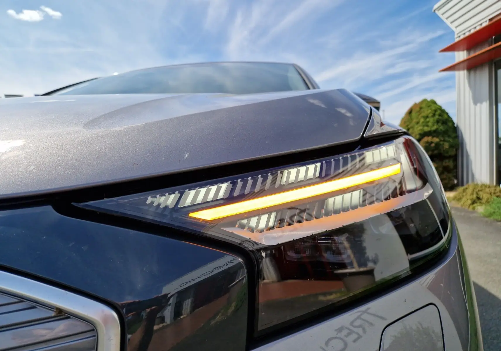 Gros plan sur le phare avant droit allumé d’un Audi Q4 E-TRON Sportback gris thyphon sous ciel bleu.