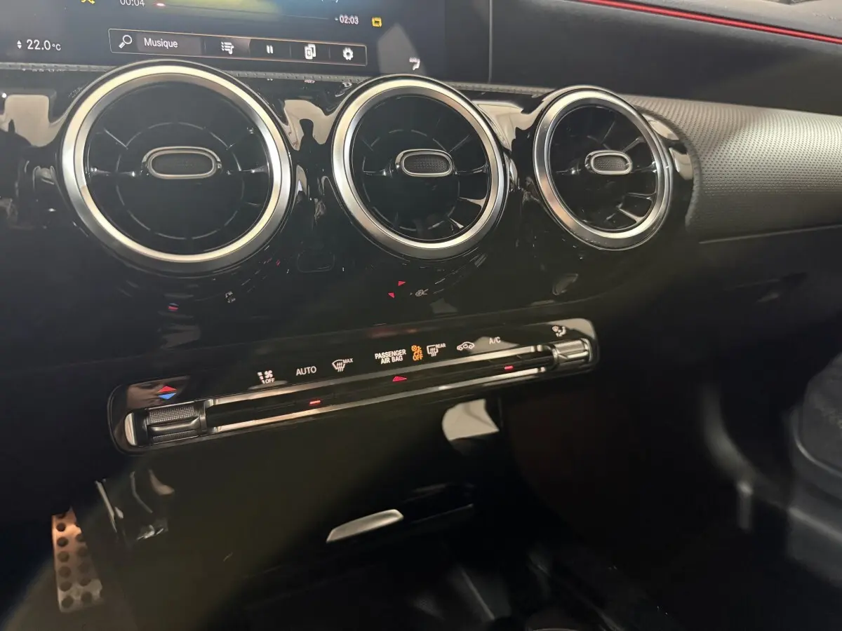 Gros plan intérieur sur la console centrale noire brillante avec aérateurs ronds chromés et bandeau tactile rouge de la Mercedes Classe A AMG Line.