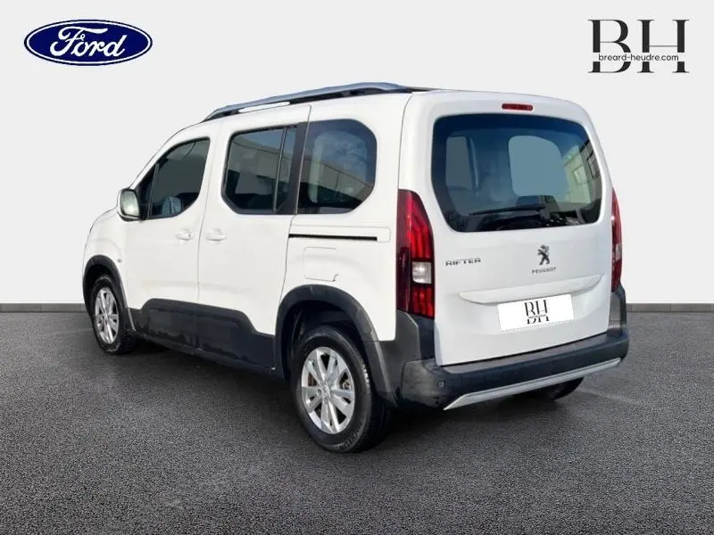 Vue 3/4 arrière droite d'un Peugeot Rifter blanc avec barres de toit et vitres teintées.