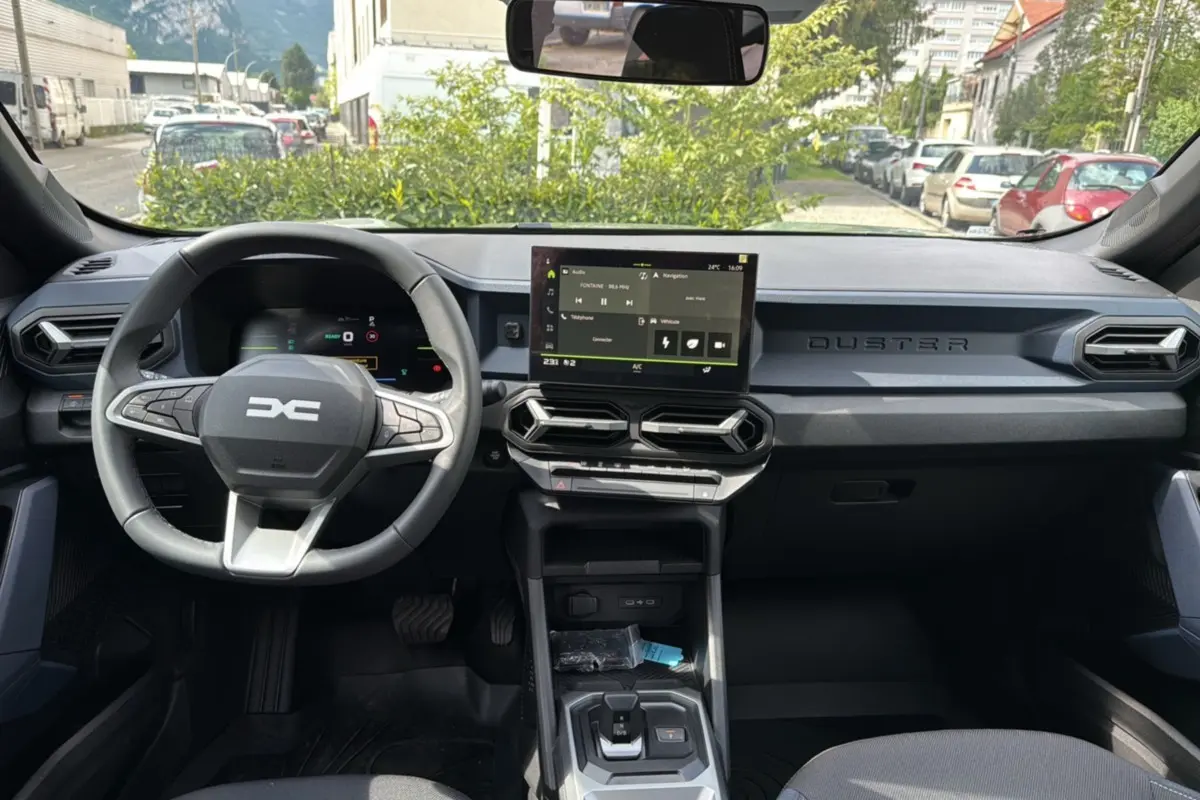 Vue intérieure avant du tableau de bord du Dacia Duster 2025, avec écran tactile central et volant multifonction noir.