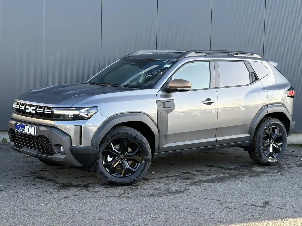 Dacia Duster gris schiste en 3/4 avant droit, avec jantes noires et coques de rétroviseur brun cuivré.