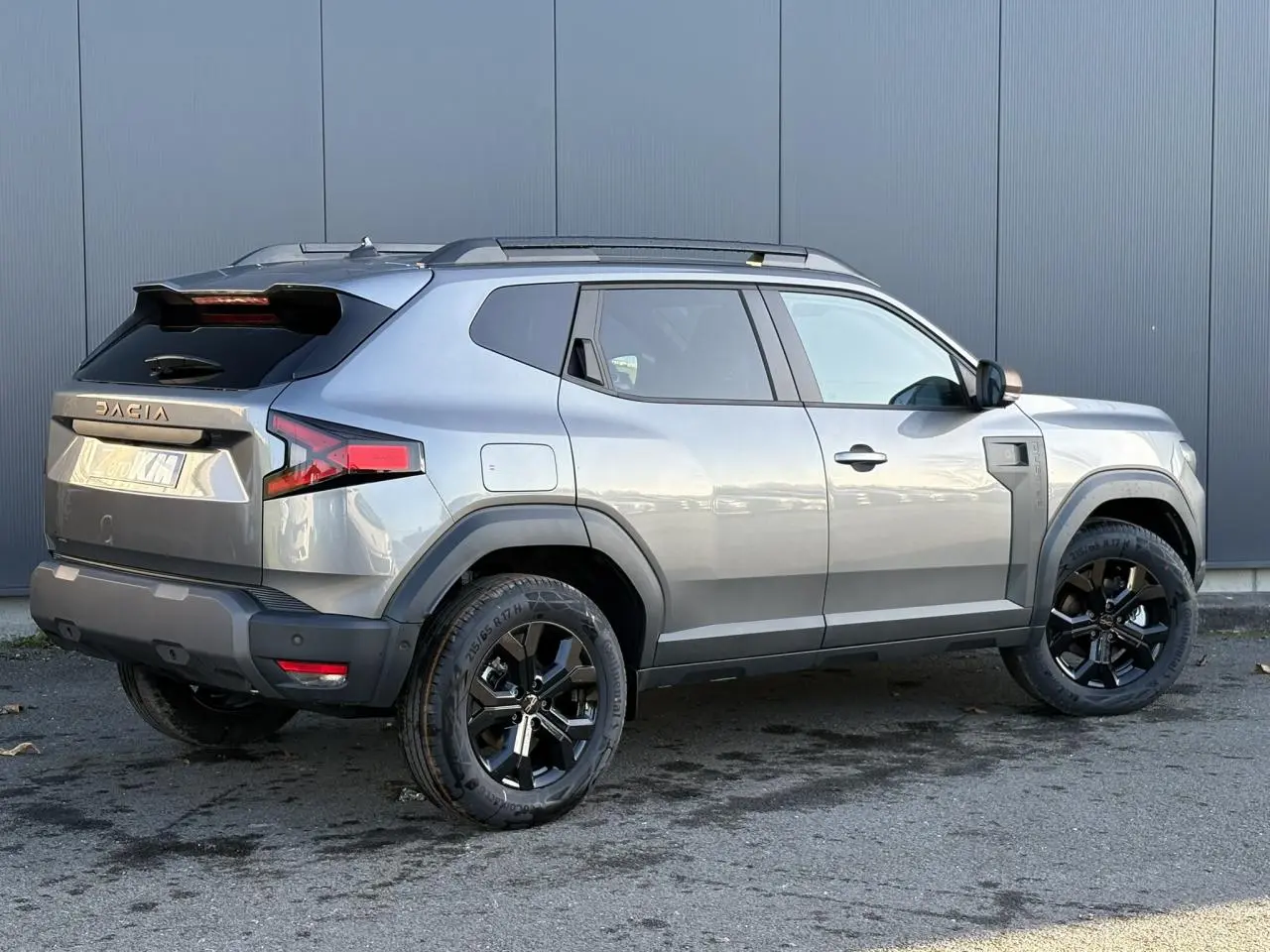 Vue 3/4 arrière droite du Dacia Duster 2025 gris schiste avec jantes noires et barres de toit noires.
