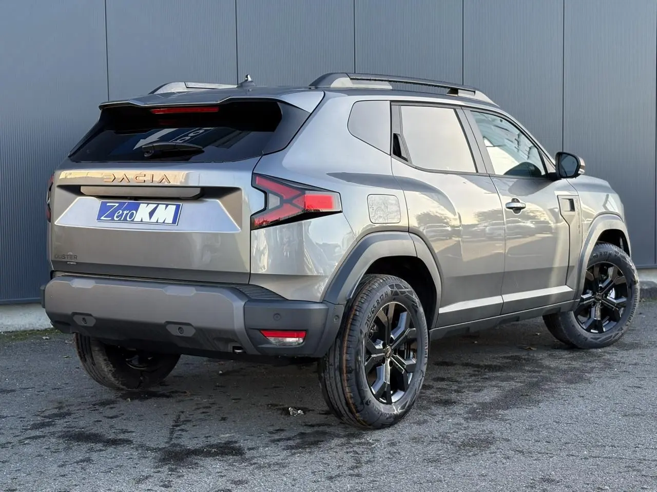 Vue 3/4 arrière droite du Dacia Duster 2025 gris schiste avec jantes noires et barres de toit longitudinales.