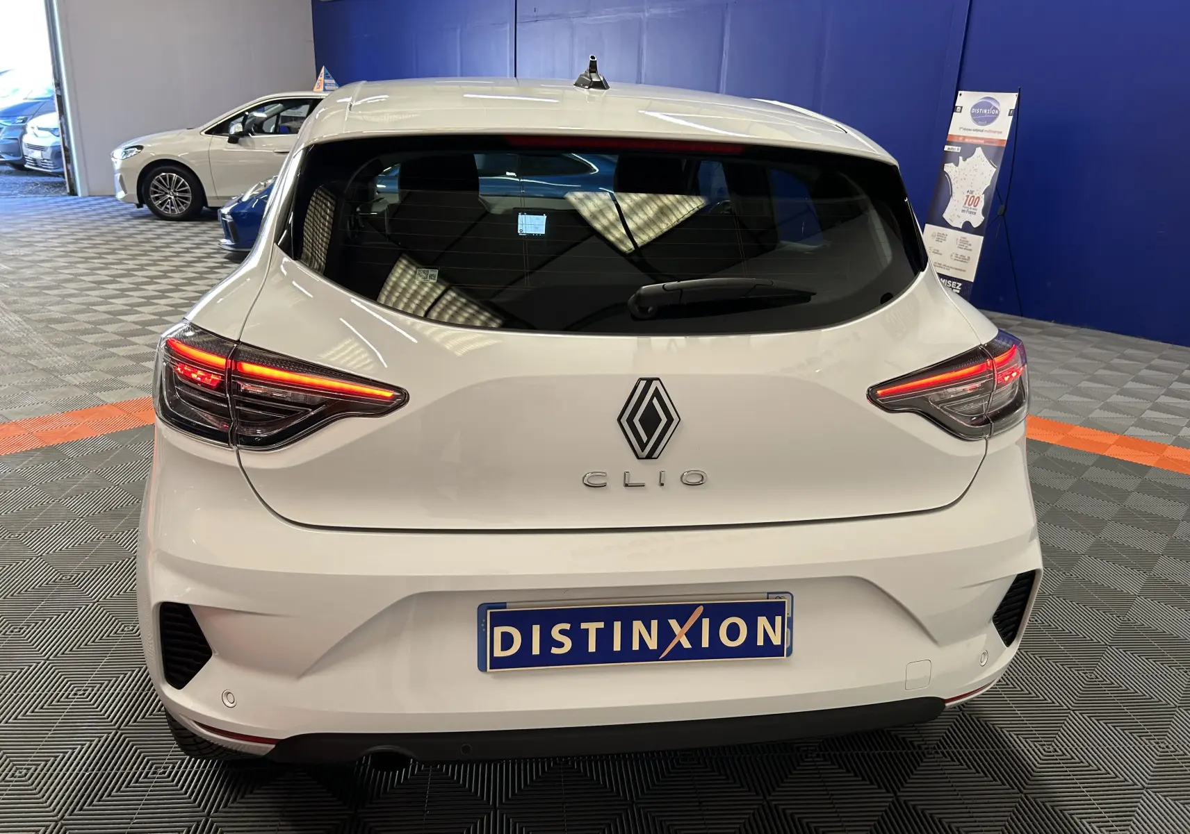 Vue arrière d'une Renault Clio V blanche avec feux arrière allumés dans un showroom au sol gris et murs bleus.