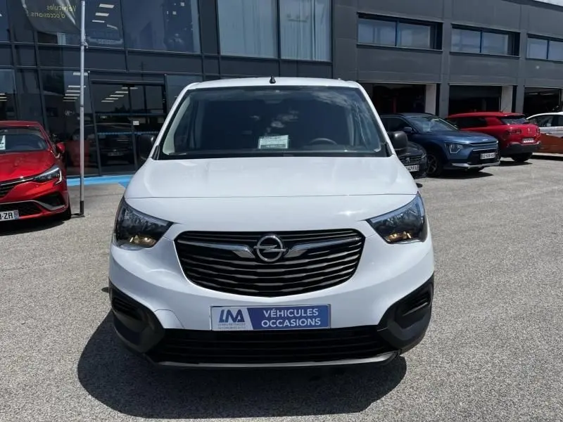 Vue avant d’un Opel Combo Cargo blanc 2024 avec calandre noire et phares allumés, stationné devant un bâtiment commercial.