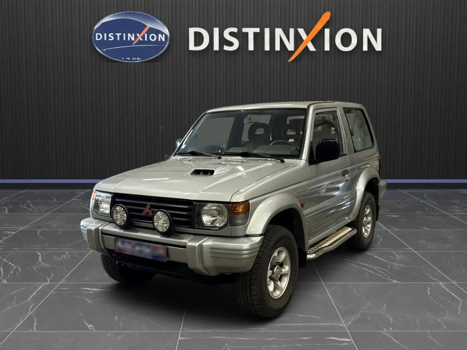 Mitsubishi Pajero Court gris aluminium vu en 3/4 avant droit, avec marchepieds et phares additionnels sur calandre.