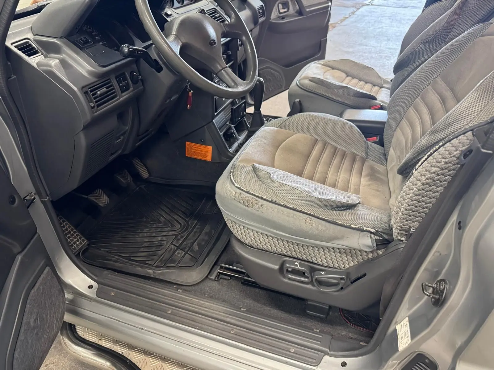 Vue intérieure côté conducteur du Mitsubishi Pajero Court gris aluminium de 1997, avec sièges tissu gris et tapis caoutchouc.
