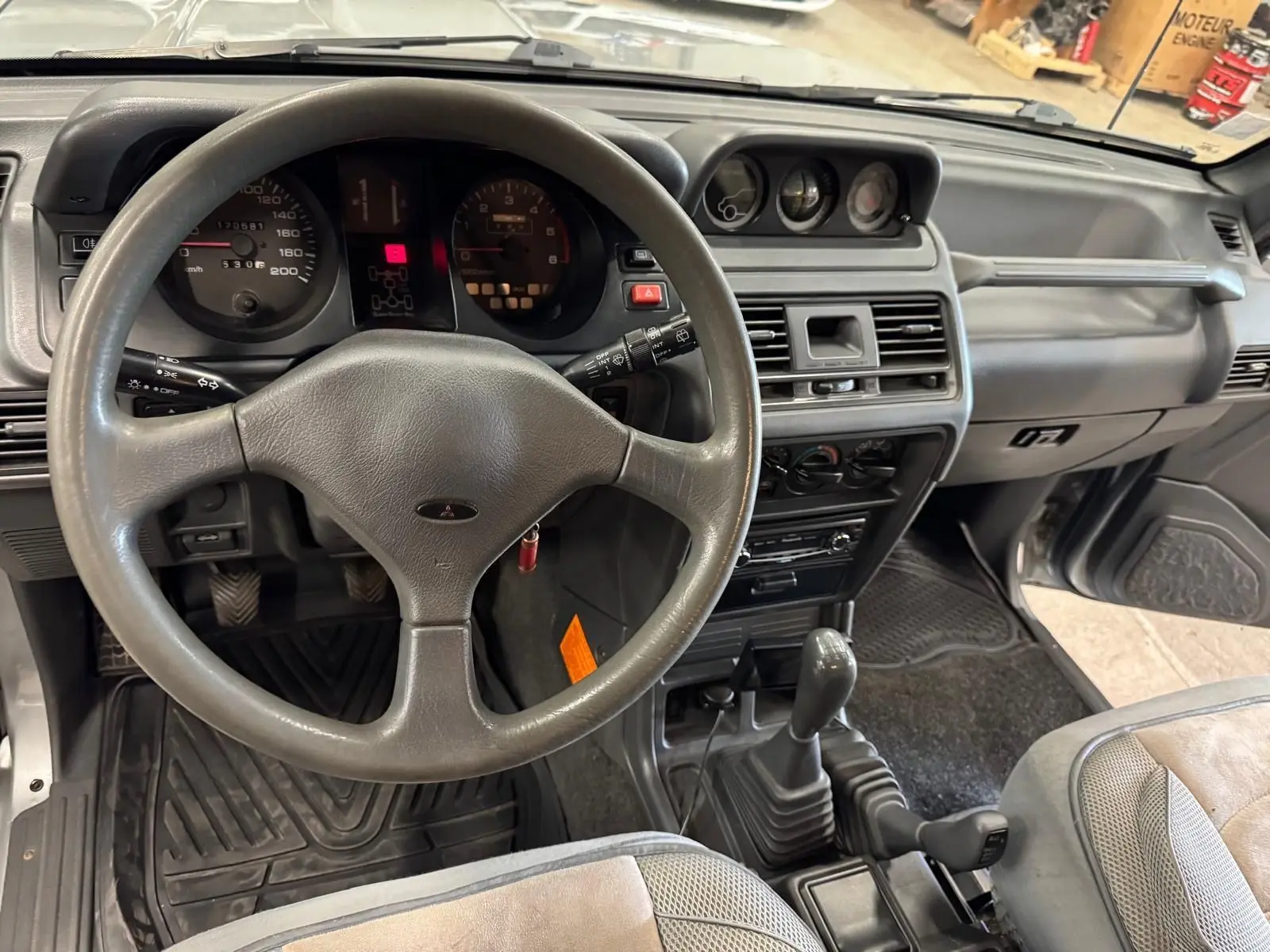 Vue intérieure du tableau de bord gris et volant du Mitsubishi Pajero Court 2.8 TD 1997 avec boîte manuelle et tapis caoutchouc.