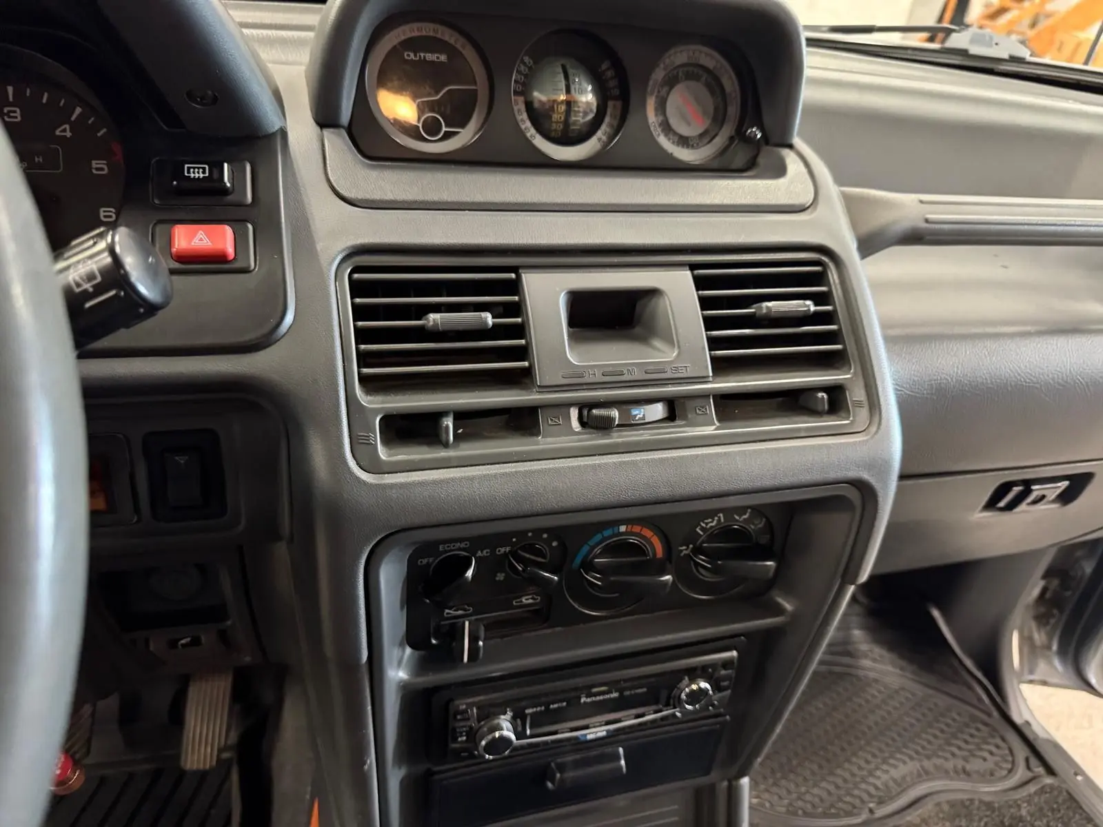 Vue rapprochée de la console centrale intérieure du Mitsubishi Pajero Court 1997, avec tableau de bord gris et commandes climatisation.