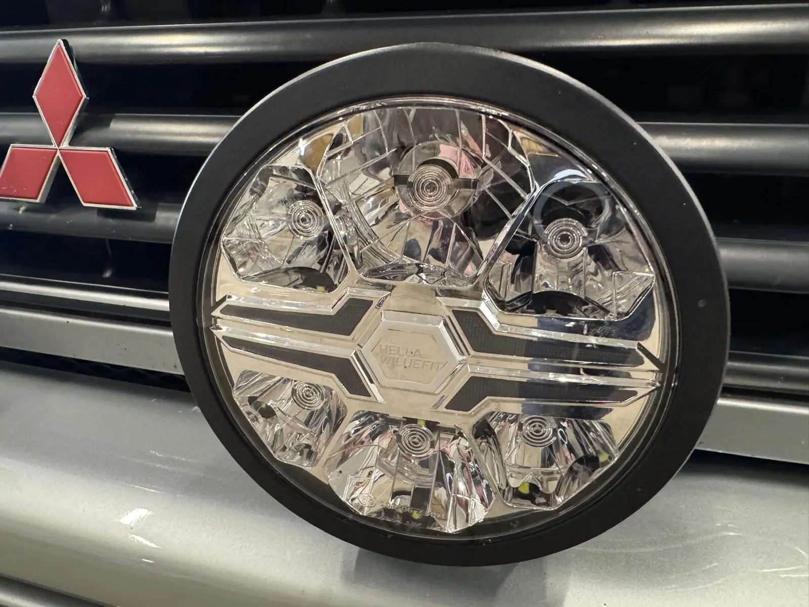 Gros plan sur le phare rond avant du Mitsubishi Pajero Court gris aluminium, avec logo rouge distinctif sur la calandre.