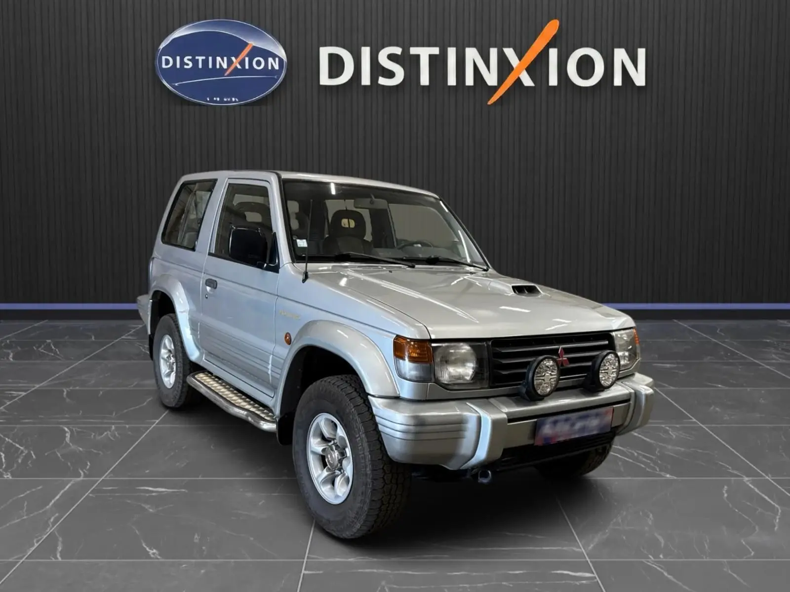 Mitsubishi Pajero Court gris aluminium vu en 3/4 avant droit avec marchepieds et deux phares additionnels frontaux.