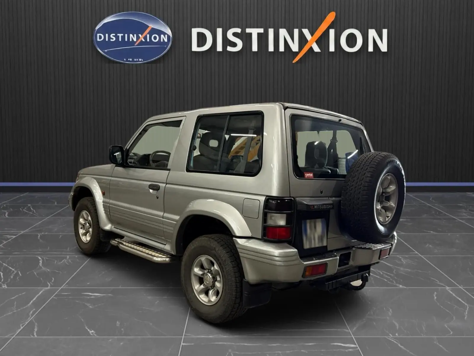 Vue 3/4 arrière droite du Mitsubishi Pajero Court gris aluminium de 1997 avec roue de secours et marchepieds visibles.