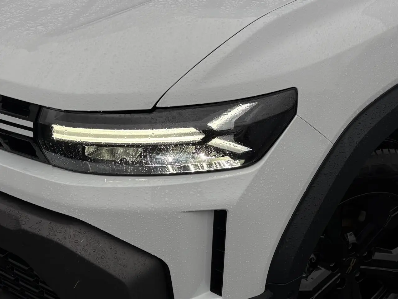 Gros plan sur le phare avant gauche du Dacia Duster blanc Glacier 2026 avec jante noire et gouttes de pluie.