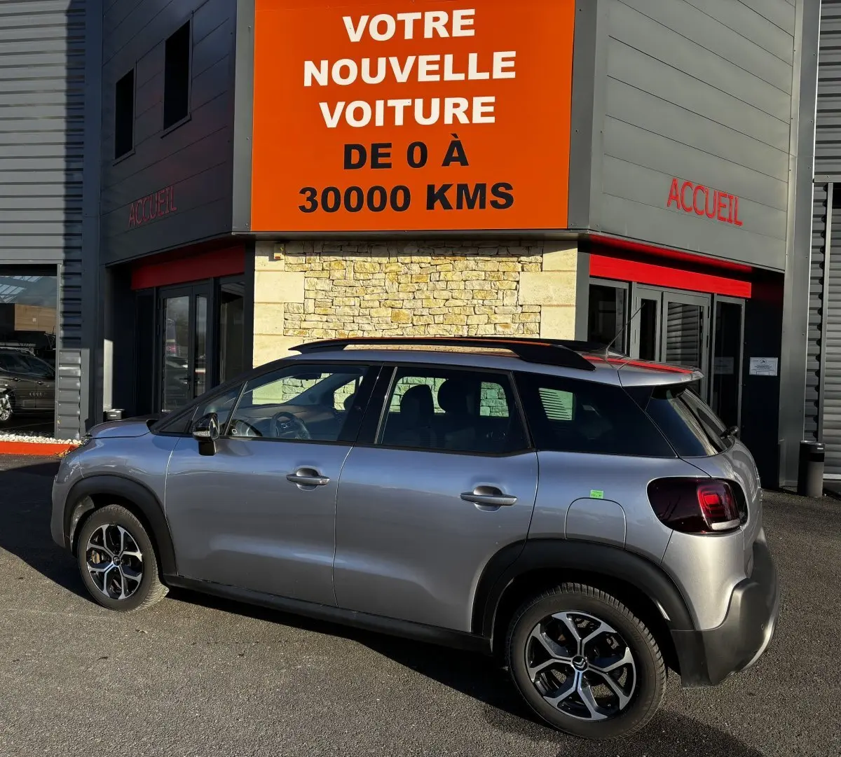 Vue latérale droite d'un Citroën C3 Aircross gris clair garé devant un bâtiment avec enseigne orange "Votre nouvelle voiture".