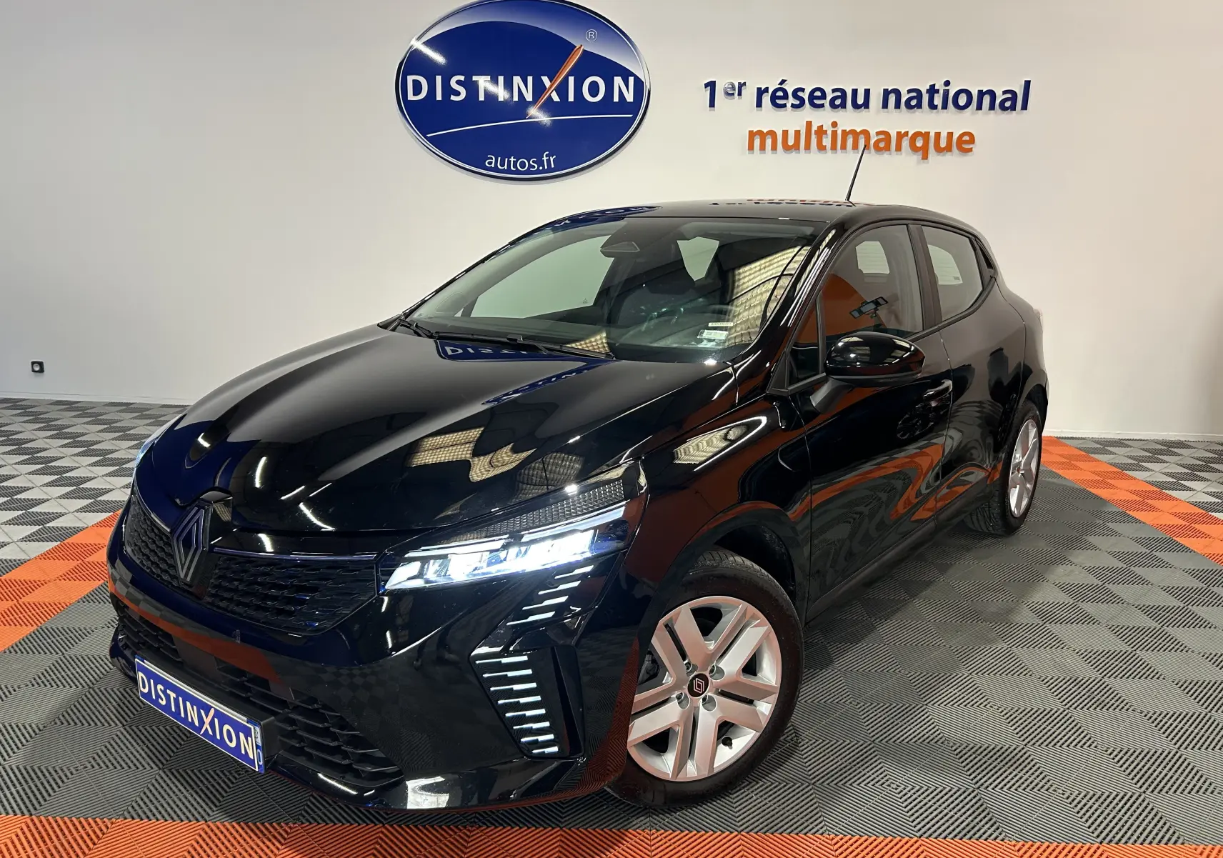 Renault Clio V noire vue en 3/4 avant droit, phares LED allumés, sur sol damier gris et orange en showroom.