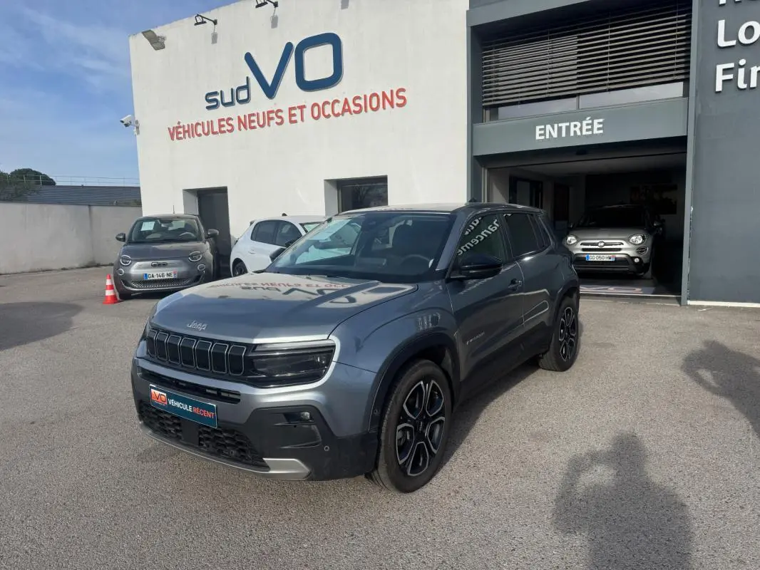 Jeep Avenger gris en 3/4 avant droit, SUV compact avec calandre noire et jantes distinctives, stationné devant un garage.