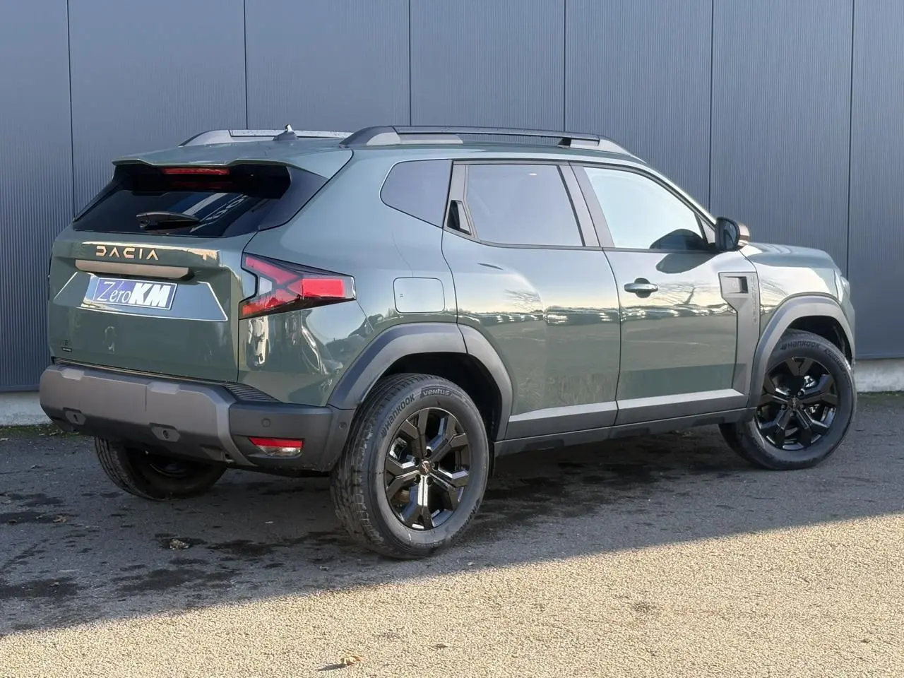 Vue 3/4 arrière droite du Dacia Duster 2025 en Kaki Lichen avec jantes noires et barres de toit grises.