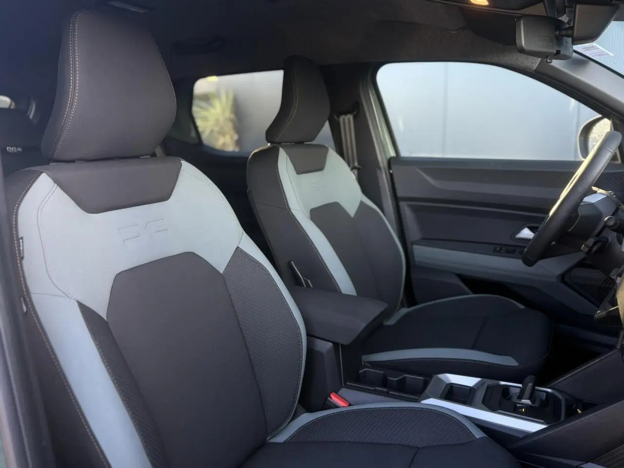Vue intérieure côté conducteur du Dacia Duster 2025, sièges bi-ton gris et noir avec accoudoir central.