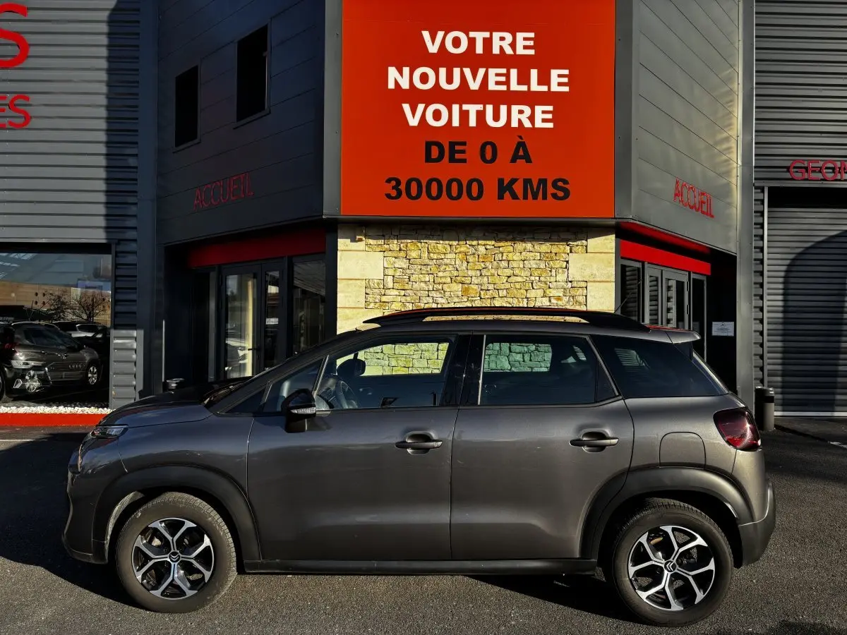 Profil latéral gauche d'un Citroën C3 Aircross gris foncé garé devant un bâtiment avec enseigne rouge.