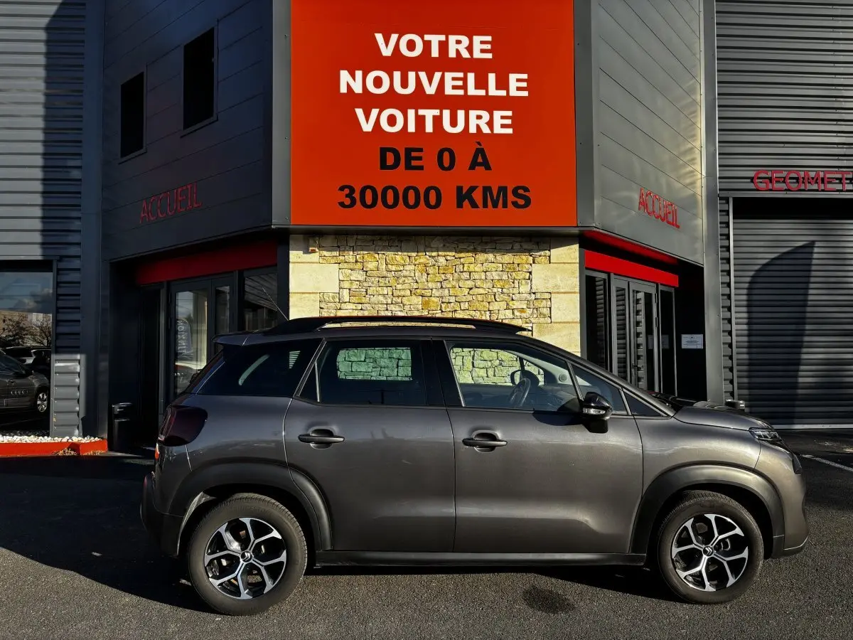 Profil côté droit d'un Citroën C3 Aircross gris foncé 2024 garé devant un bâtiment avec panneau rouge "Votre nouvelle voiture".