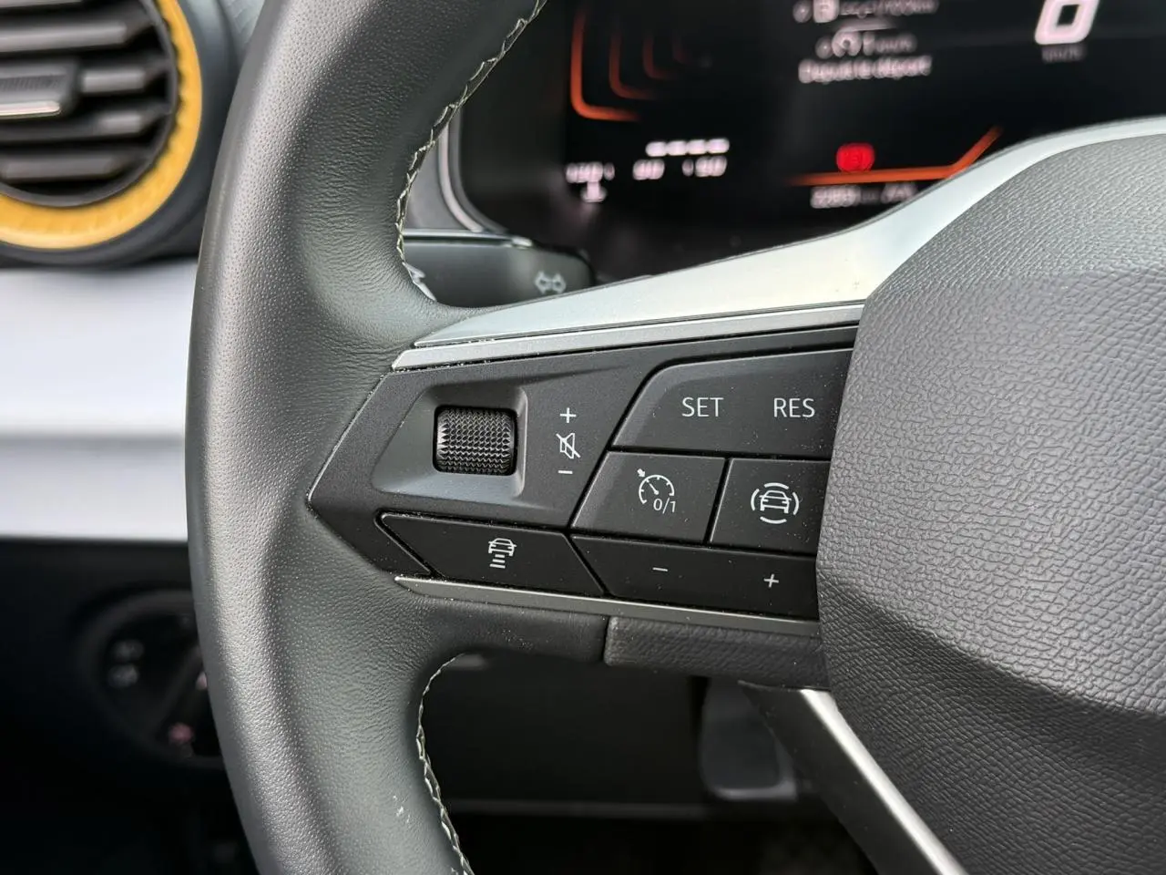 Gros plan sur les commandes gauche du volant noir du SEAT Arona 2025, avec tableau de bord digital en arrière-plan.