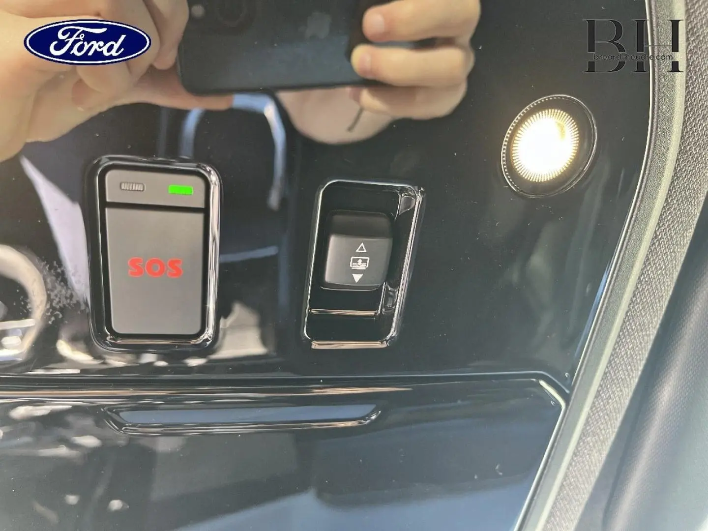 Panneau de commandes intérieur noir brillant avec bouton SOS rouge et éclairage, reflet de mains tenant un téléphone.