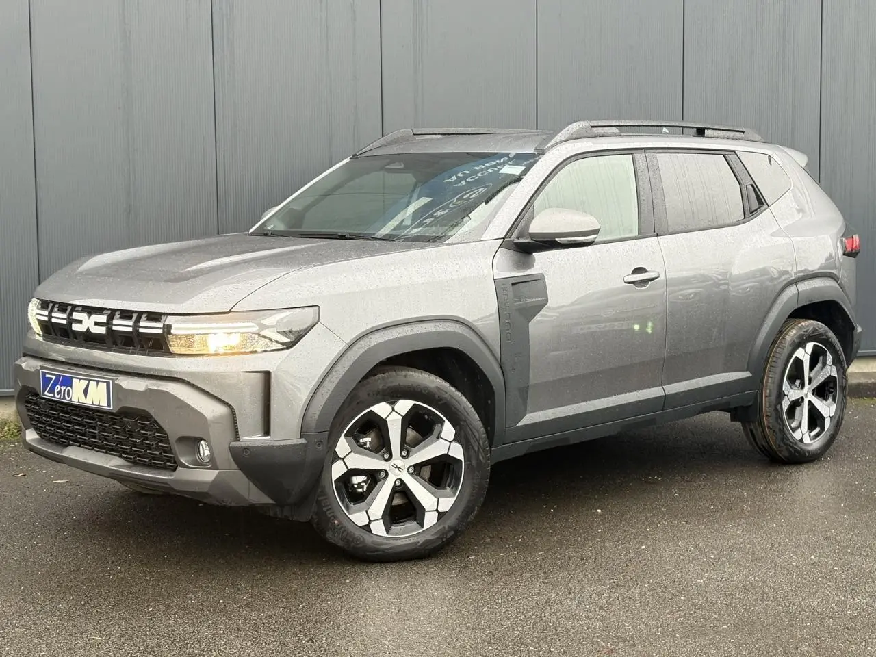 SUV Dacia Duster gris schiste en 3/4 avant droit, avec jantes alliage diamantées et barres de toit noires.