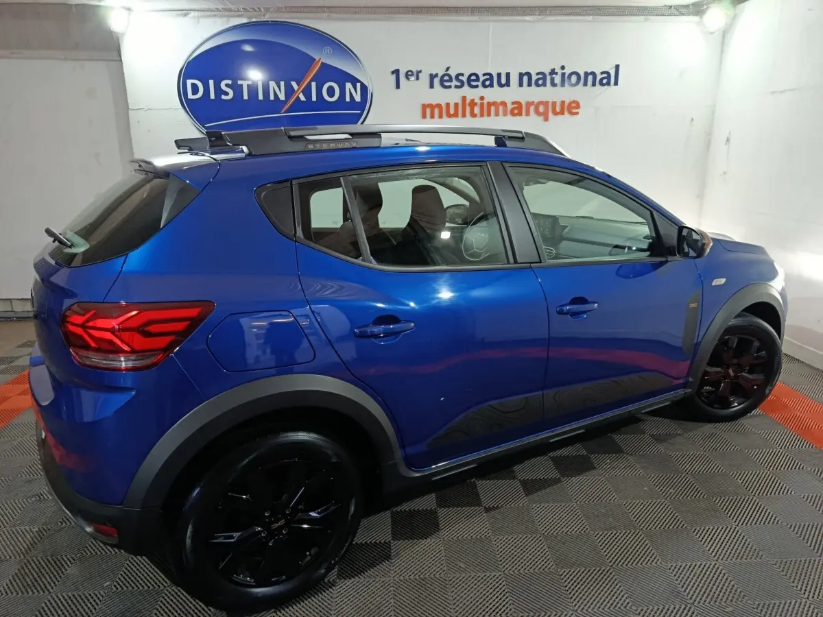 Vue côté droit d'une Dacia Sandero Extreme+ TCe 110 bleu 2024 avec jantes noires et barres de toit assorties.