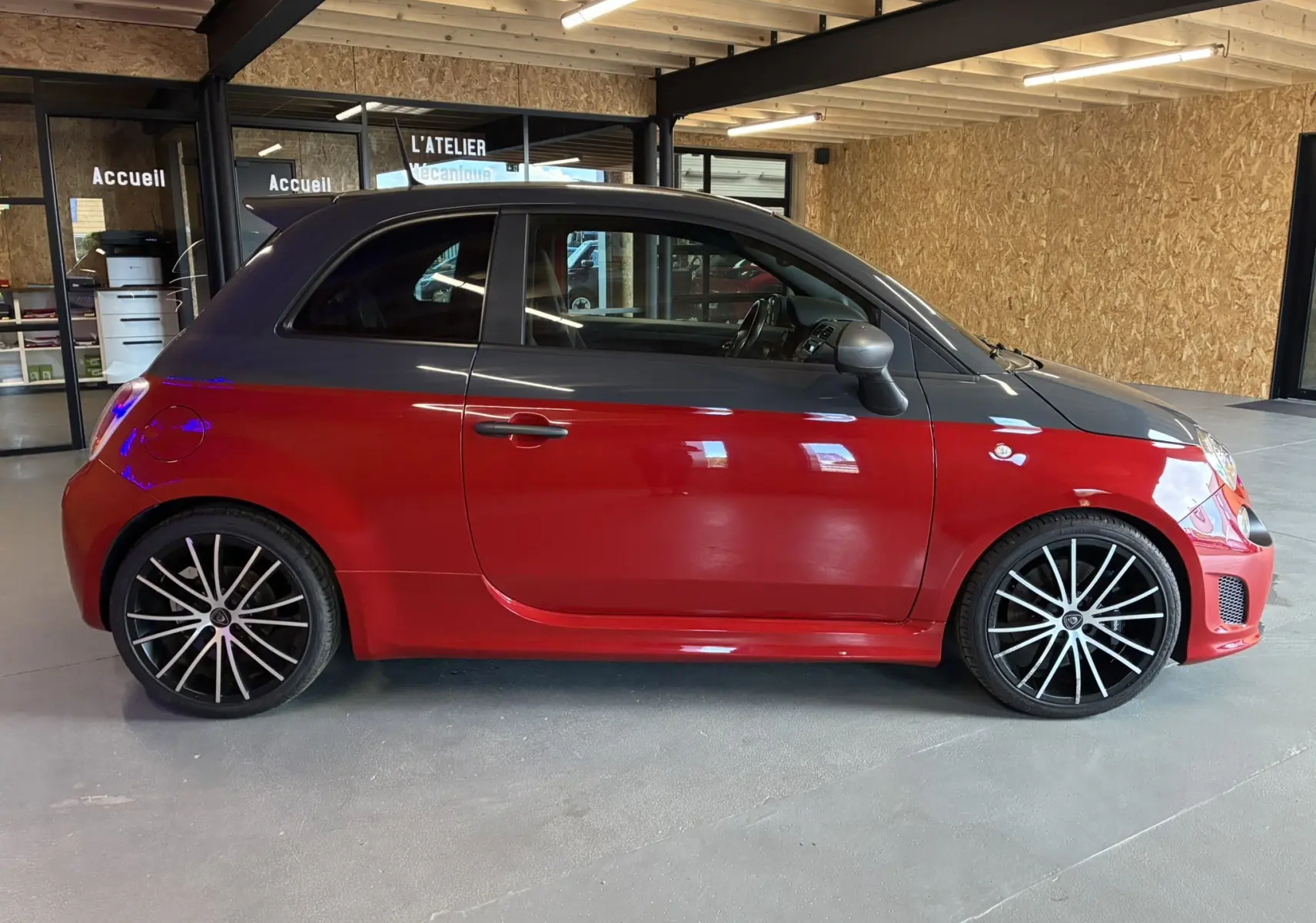 Profil droit d'une Abarth 500 Competizione 2016 rouge et gris avec jantes noires à rayons fins, en intérieur d'atelier.
