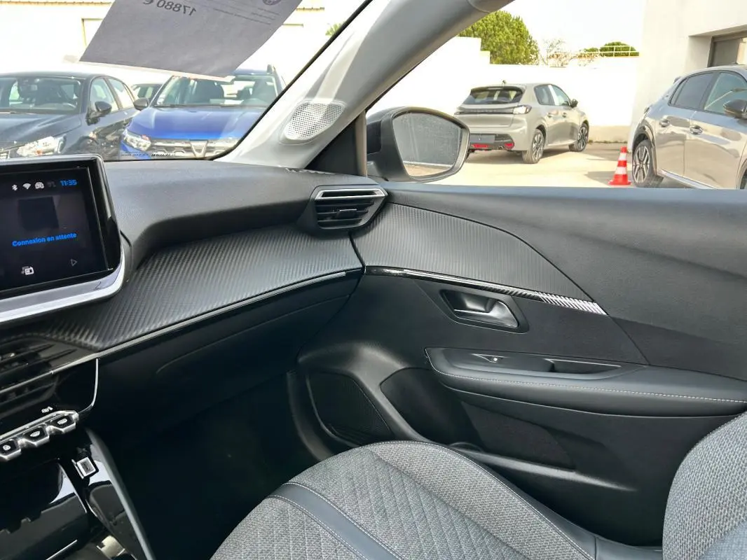 Vue intérieure côté passager du Peugeot 208 Hybrid 110 e-DCS6 Allure 2024, avec tableau de bord noir et sièges gris.
