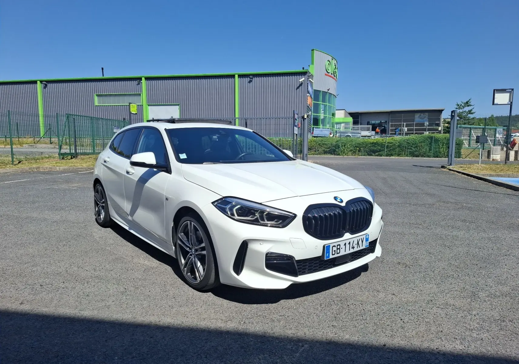 BMW Série 1 118D M Sport blanc vue 3/4 avant droit, jantes alliage 18 pouces et toit ouvrant visible.