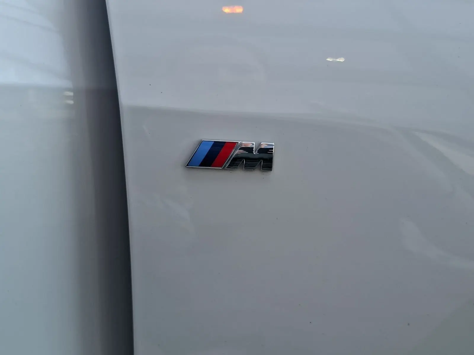 Détail du logo M sur la carrosserie blanche d'une BMW Série 1 118D M Sport, vue rapprochée côté gauche.