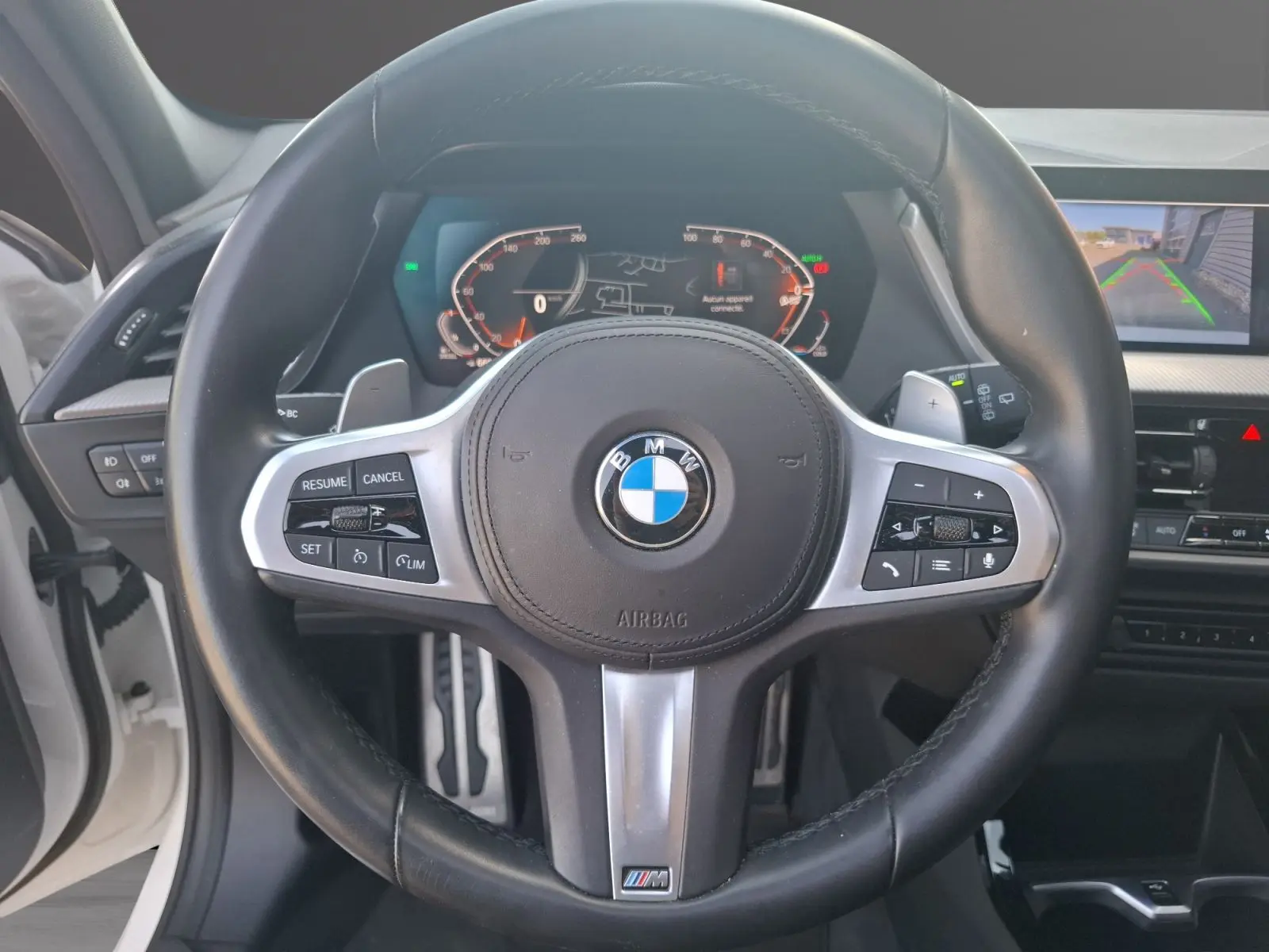 Vue intérieure centrée sur le volant cuir M Sport d'une BMW Série 1 blanche avec tableau de bord numérique et caméra de recul.