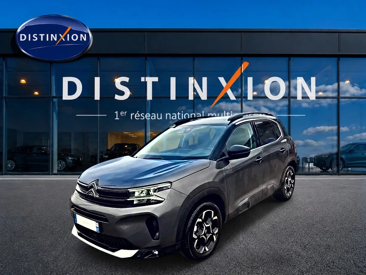 Citroën C5 Aircross gris en 3/4 avant droit, avec jantes alu et barres de toit visibles devant showroom Distinxion.