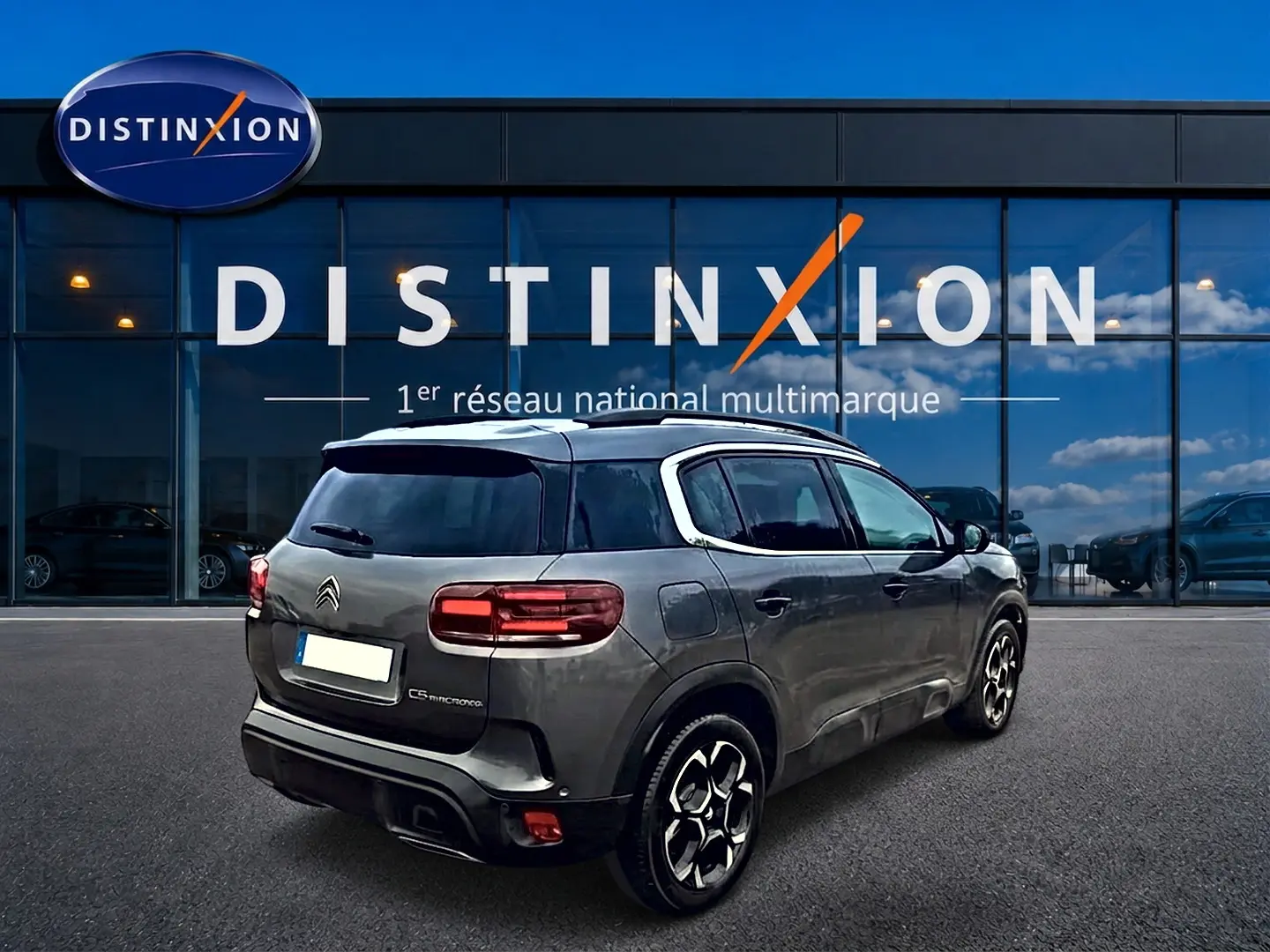 Vue 3/4 arrière droite d'un Citroën C5 Aircross gris 2025 avec barres de toit et jantes alu devant un showroom Distinxion.