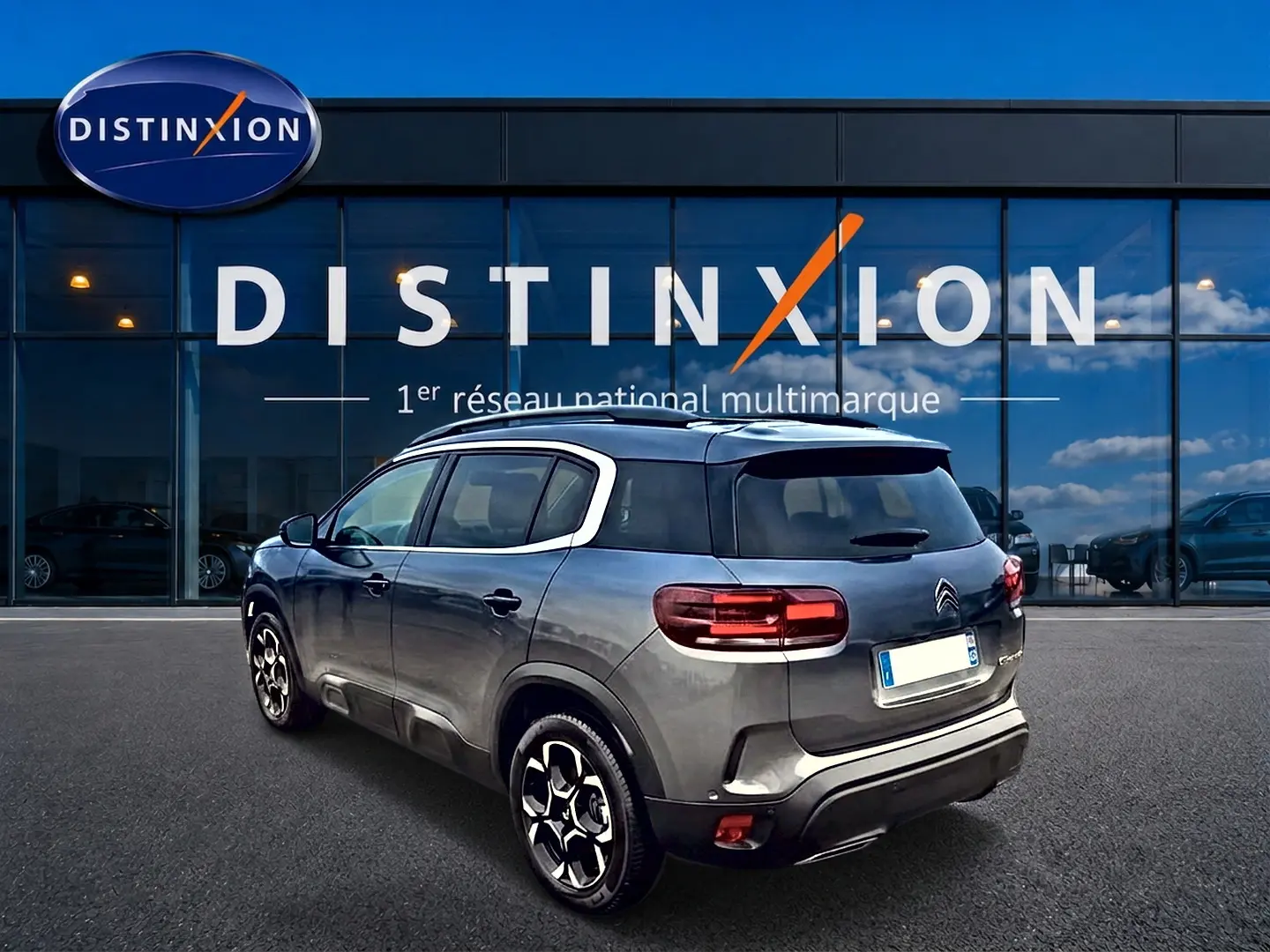 Vue 3/4 arrière droite d'un Citroën C5 Aircross gris 2025 avec jantes alu et barres de toit devant un showroom Distinxion.