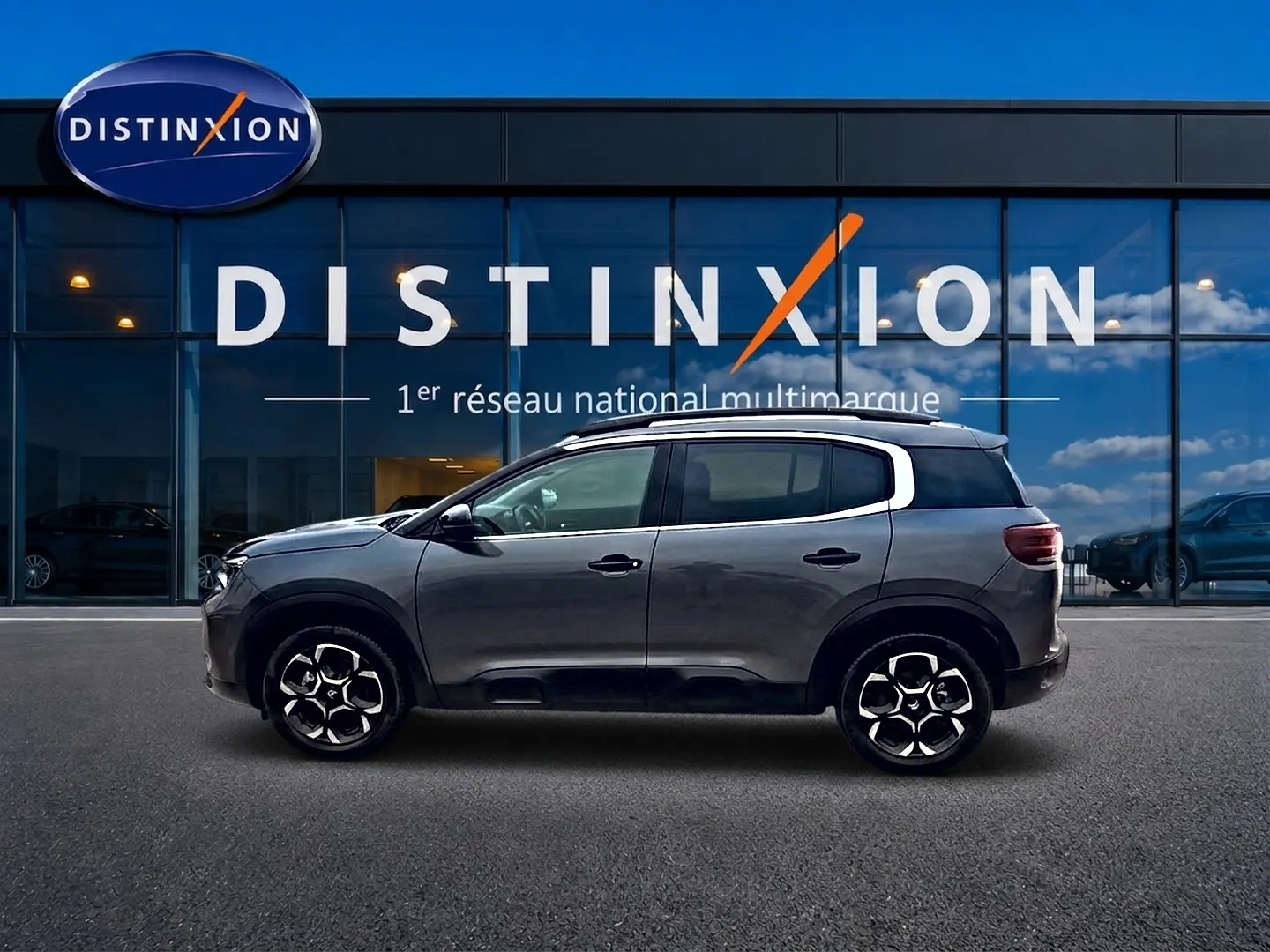 Profil côté gauche du Citroën C5 Aircross gris 2025, avec jantes alu et barres de toit visibles devant une vitrine Distinxion.
