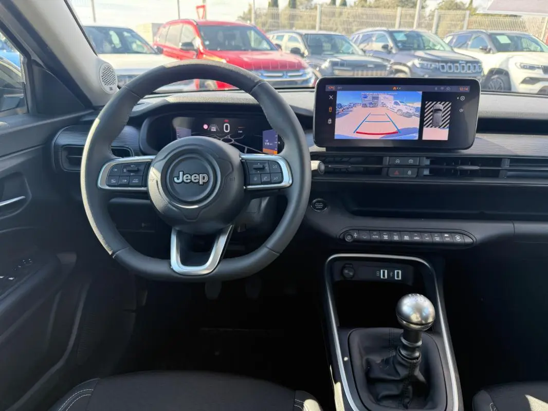 Vue intérieure frontale du tableau de bord du Jeep Avenger blanc 2025 avec volant multifonction et écran tactile affichant la caméra de recul.