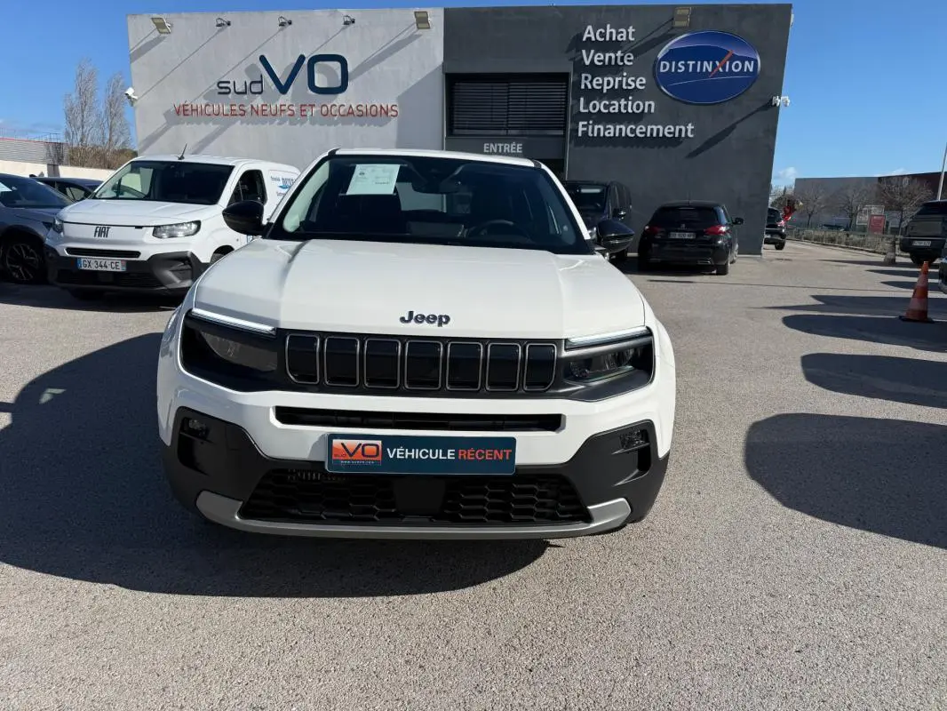 Vue avant d'un Jeep Avenger blanc 2025 avec calandre noire et phares LED, stationné devant un concessionnaire.