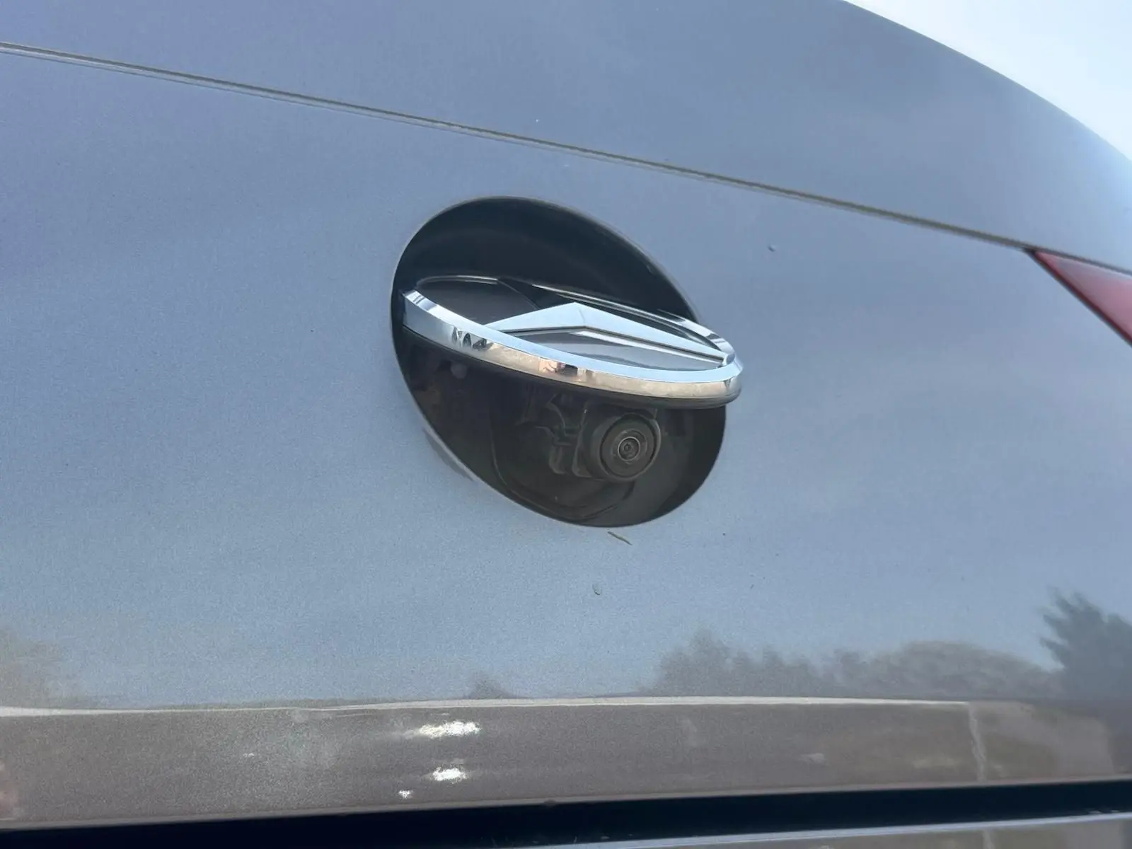 Gros plan sur la caméra de recul intégrée sous le logo chromé à l'arrière d'une Mercedes CLA 180 D gris montagne.