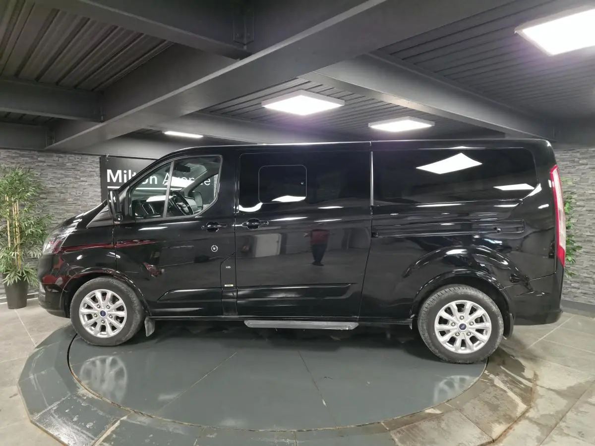 Vue de profil côté gauche d’un Ford Transit Fourgon Custom noir métallisé avec jantes alliage 17 pouces en intérieur showroom.