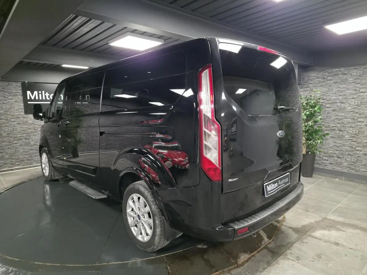 Vue 3/4 arrière droite du Ford Transit Fourgon Custom noir, avec jantes alliage et hayon vitré en intérieur showroom.