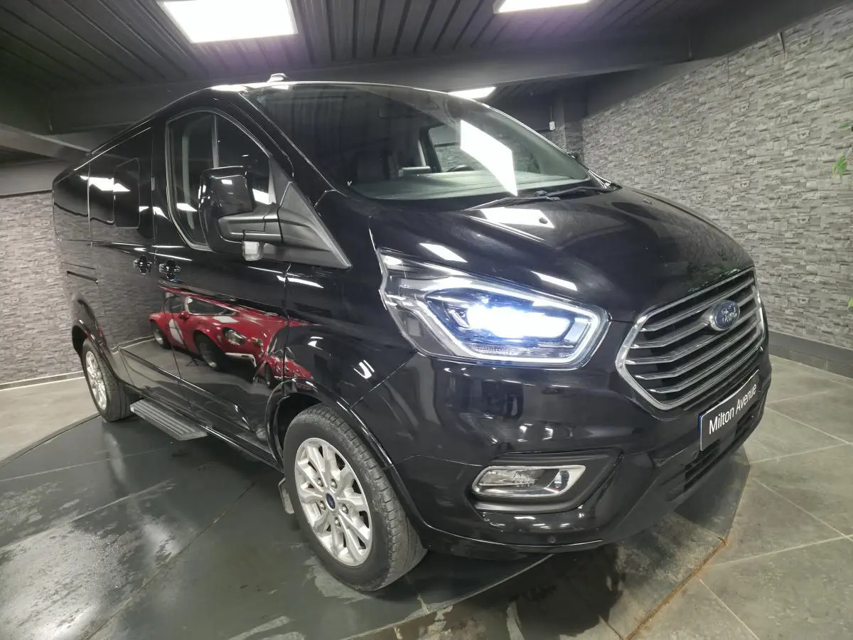 Ford Transit Fourgon Custom noir métallisé vu en 3/4 avant droit avec phares xénon allumés dans un garage.