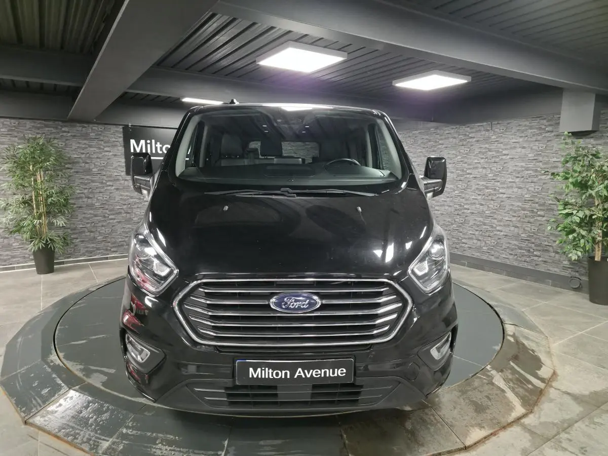 Vue frontale du Ford Transit Fourgon Custom noir, avec calandre chromée et phares allumés en showroom.